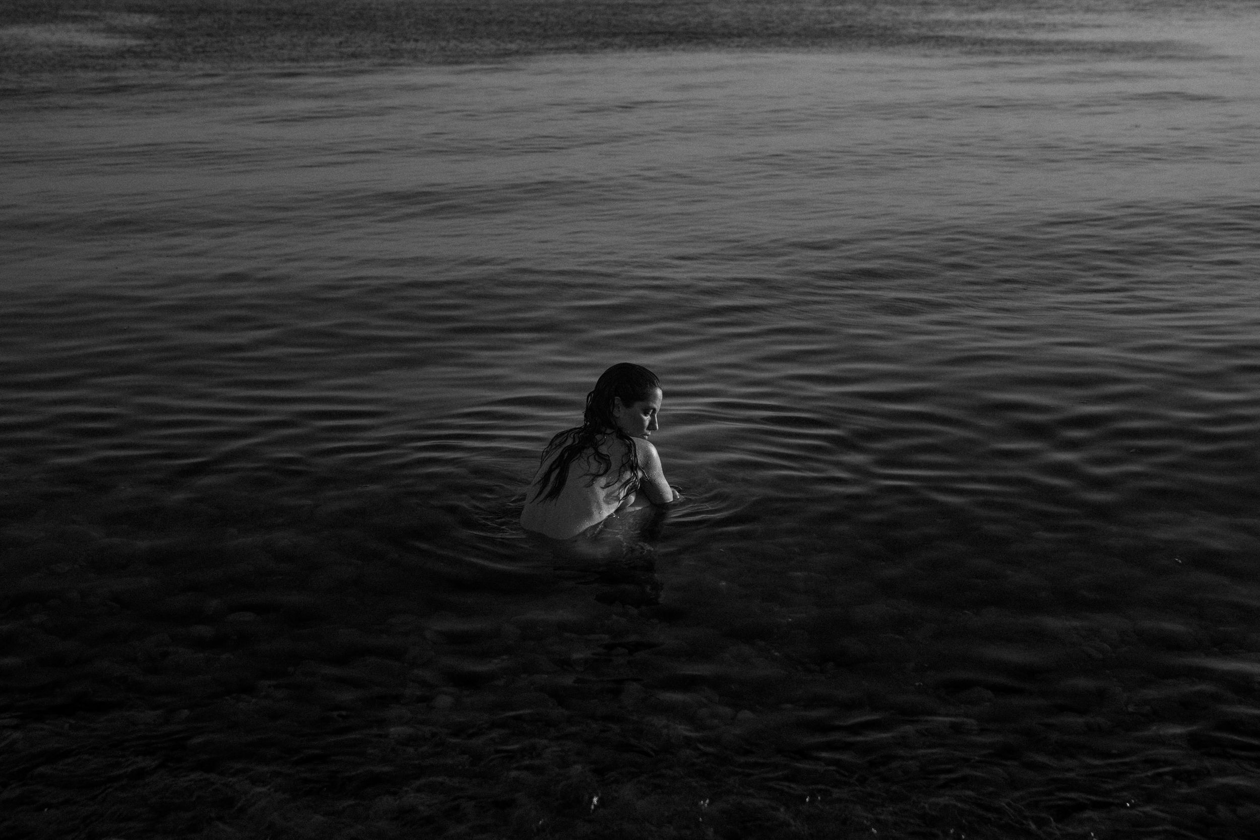 Black and white photo of a woman standing waist-deep in calm water, looking contemplative.