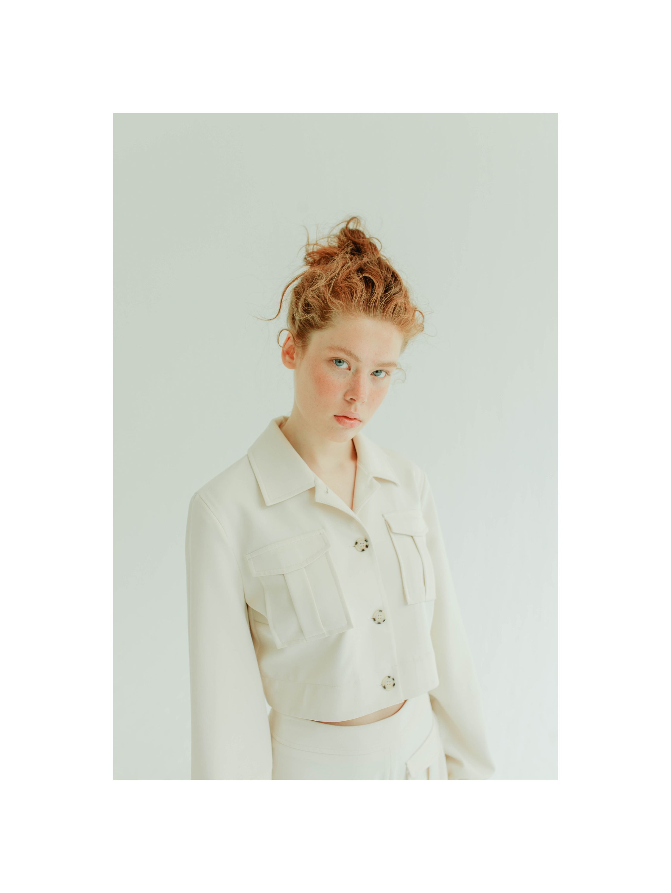 Young woman with red hair in a bun wearing a cream cropped jacket with gold buttons against white background.
