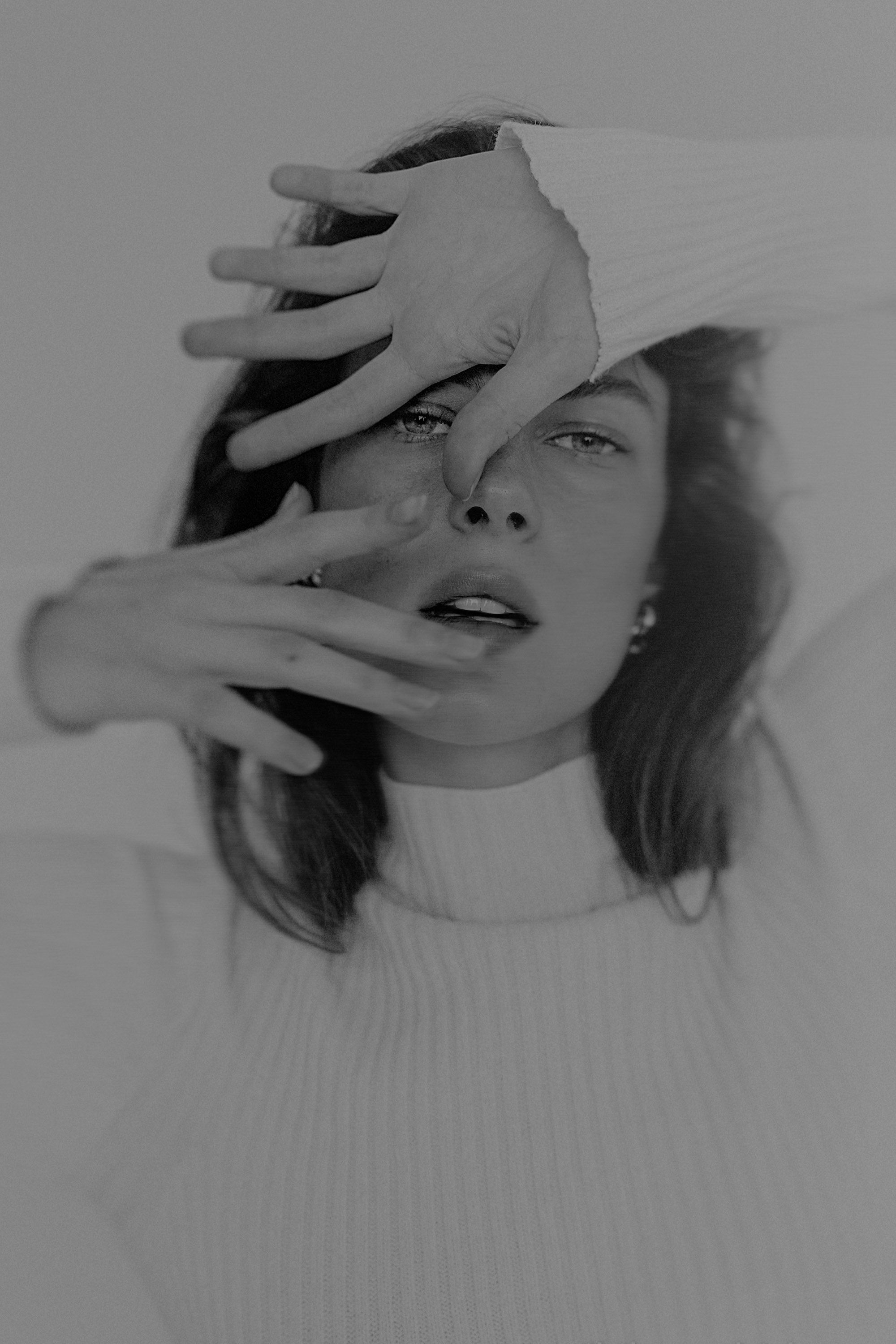 Black and white portrait of a woman covering her face with hands, wearing a white turtleneck sweater.