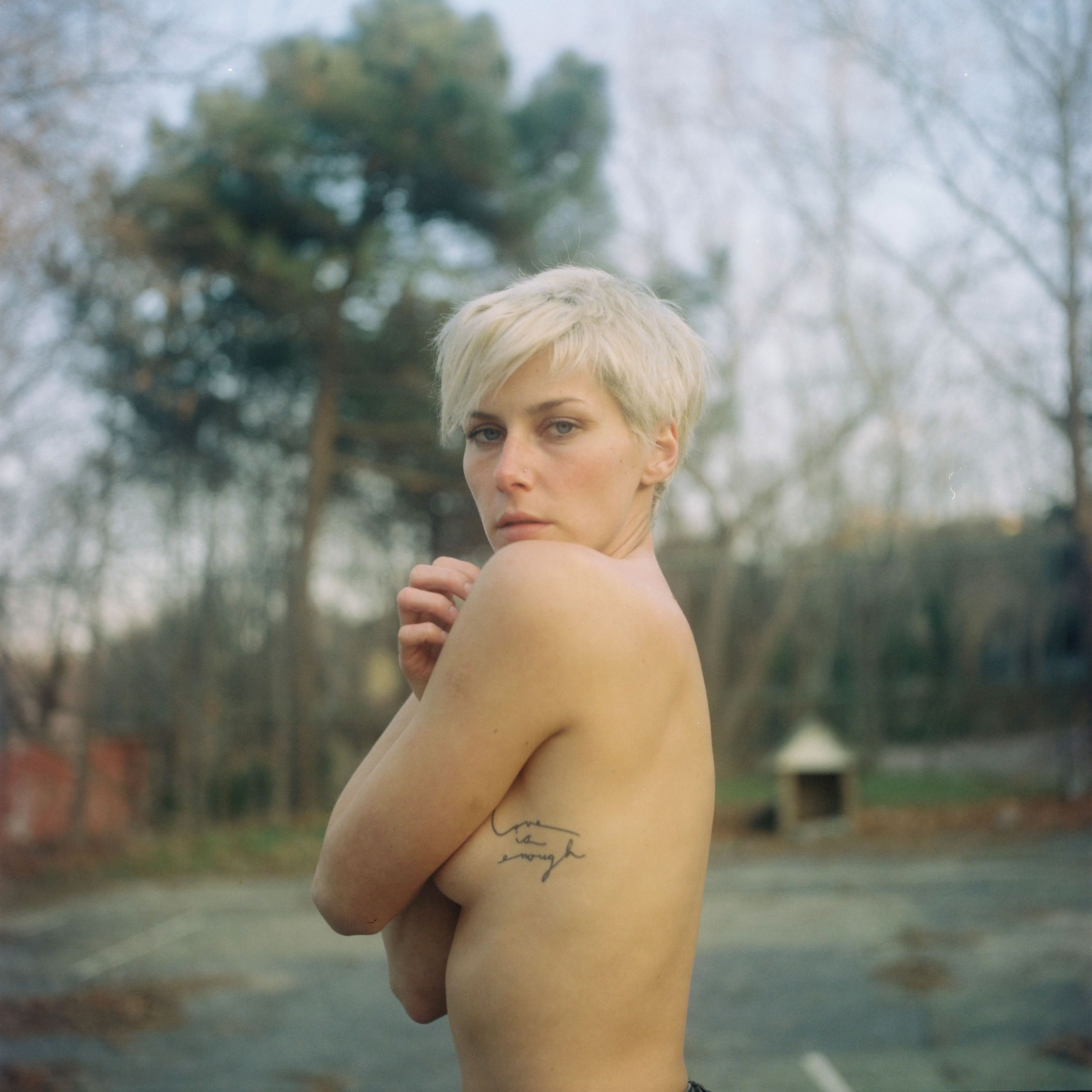 A blonde woman with a short pixie cut and arm tattoo poses outdoors near trees in natural light. Yashica Mat 124 Analogue Photography.