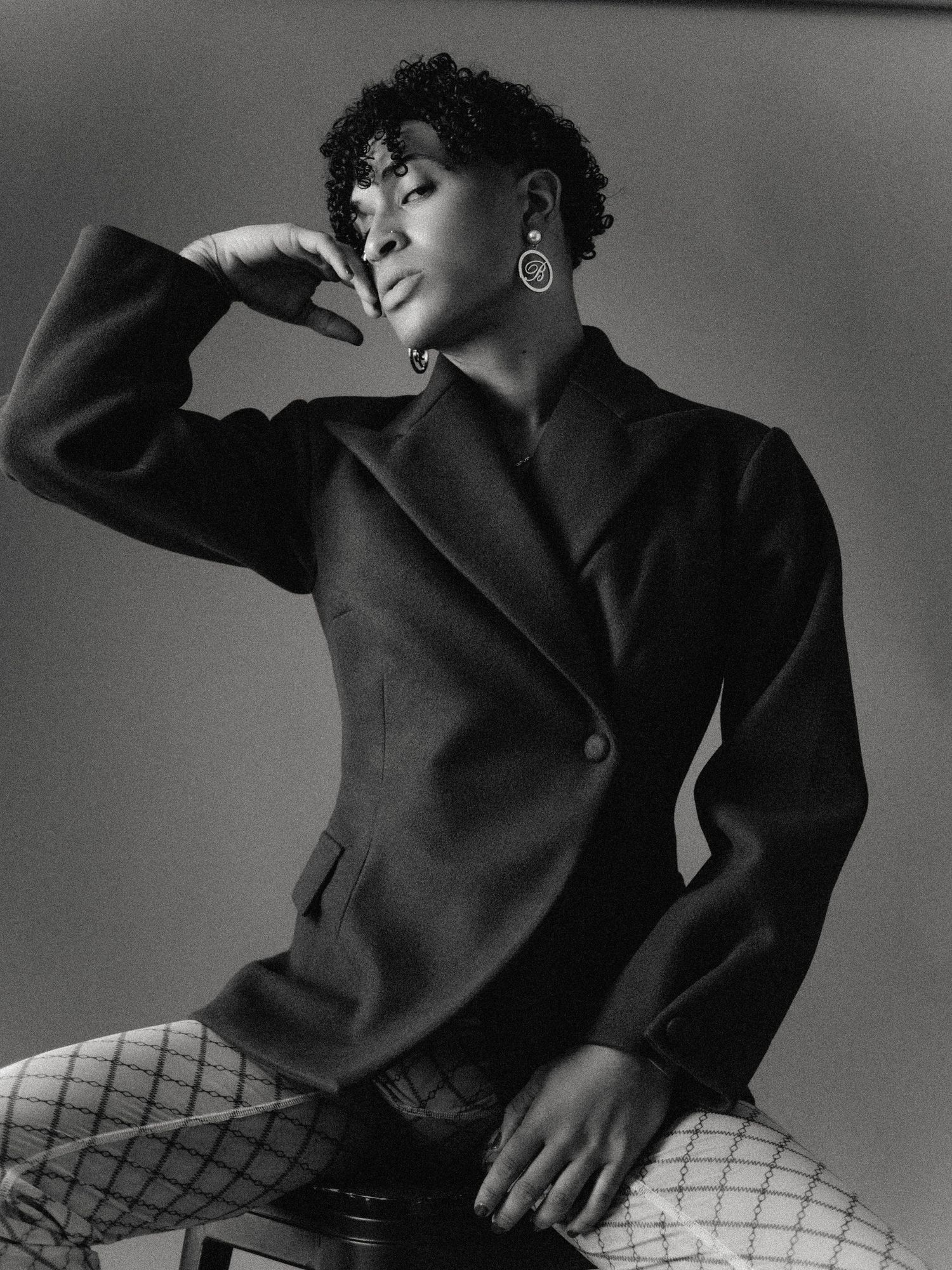 Black and white portrait of a person in a blazer and fishnet tights, posing thoughtfully with hand raised.
