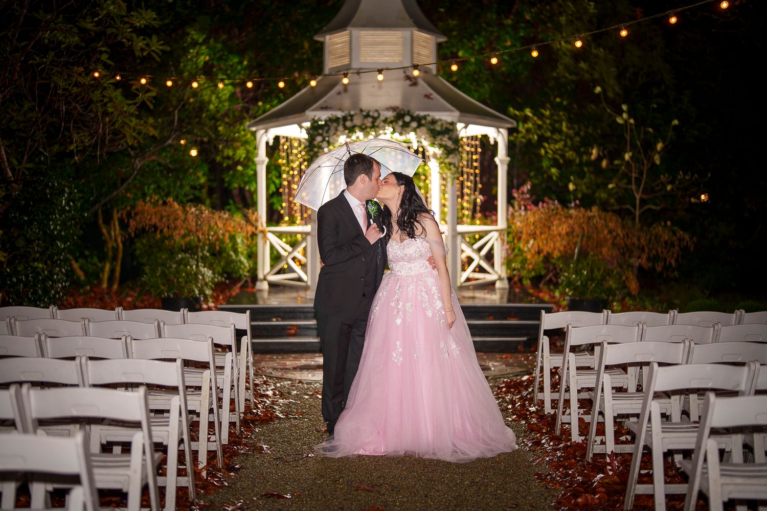 Bride in pink gown and groom kiss at night beside lit gazebo with string lights at wedding venue.