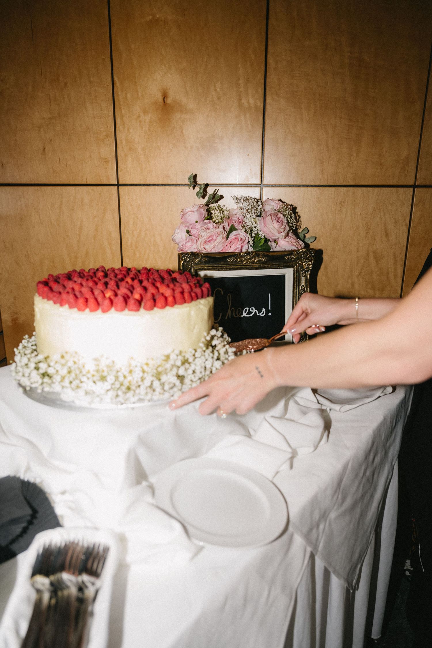 Bride arranging a white wedding cake topped with fresh strawberries and small white flowers on a draped table.
