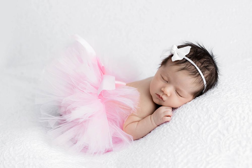 Sleeping newborn baby girl wearing a pink tutu skirt and white bow headband on a white blanket.
