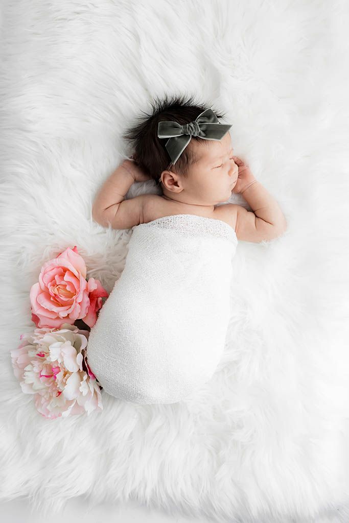 Sleeping newborn baby girl swaddled in white wrap with pink flowers on fluffy white fur backdrop.