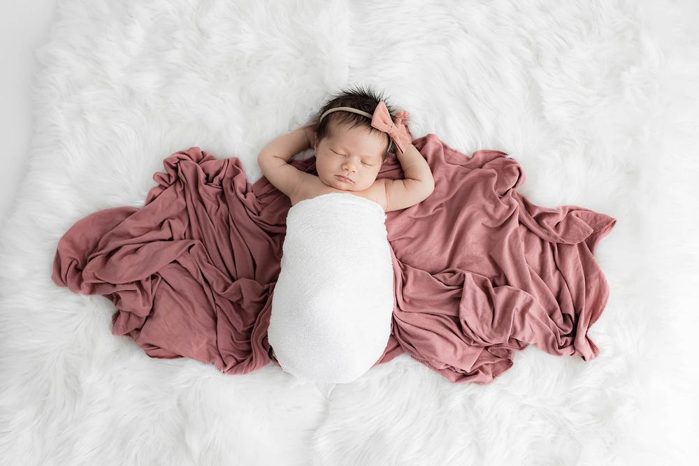 Peaceful newborn baby girl wrapped in white swaddle on dusty rose blanket and white fur rug.