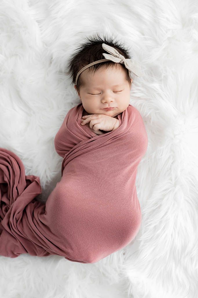 Newborn baby girl sleeping peacefully swaddled in dusty pink wrap with bow headband on white fur.
