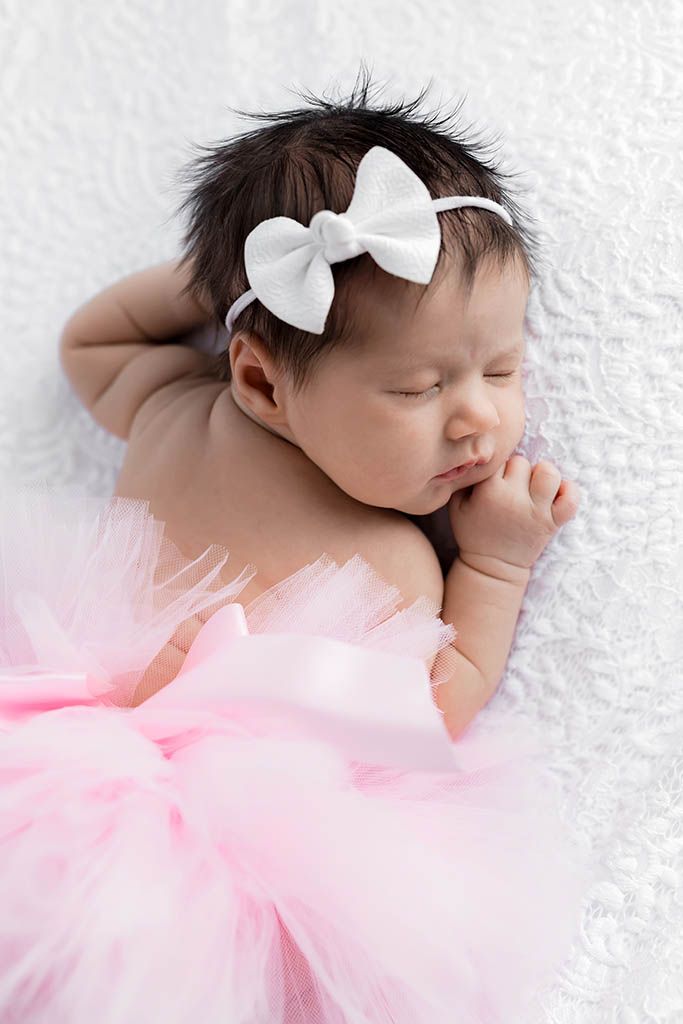 Sleeping newborn baby girl in pink tutu skirt with white bow headband on soft white background.
