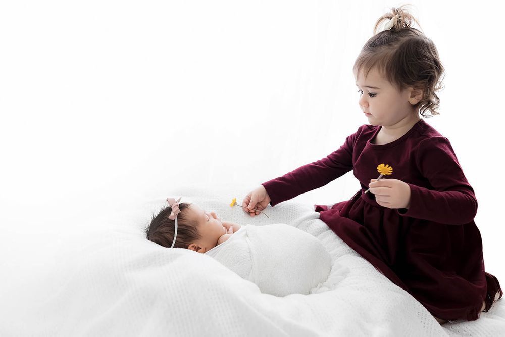 Sweet toddler girl in burgundy dress offering yellow flower to sleeping newborn sibling in white wrap.