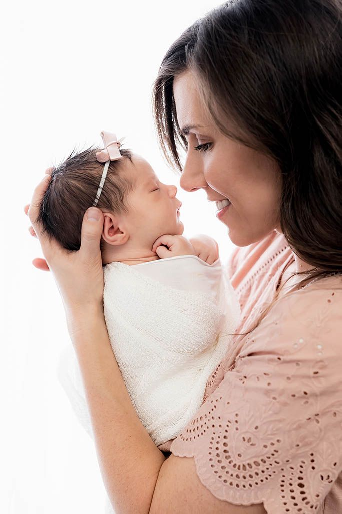 Smiling dark-haired mother holds swaddled newborn baby girl wearing pink bow headband against white background.