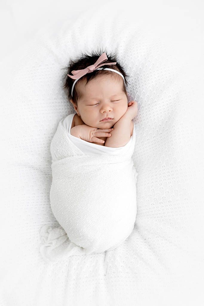 Sleeping newborn baby girl swaddled in white wrap with pink bow headband on white blanket.