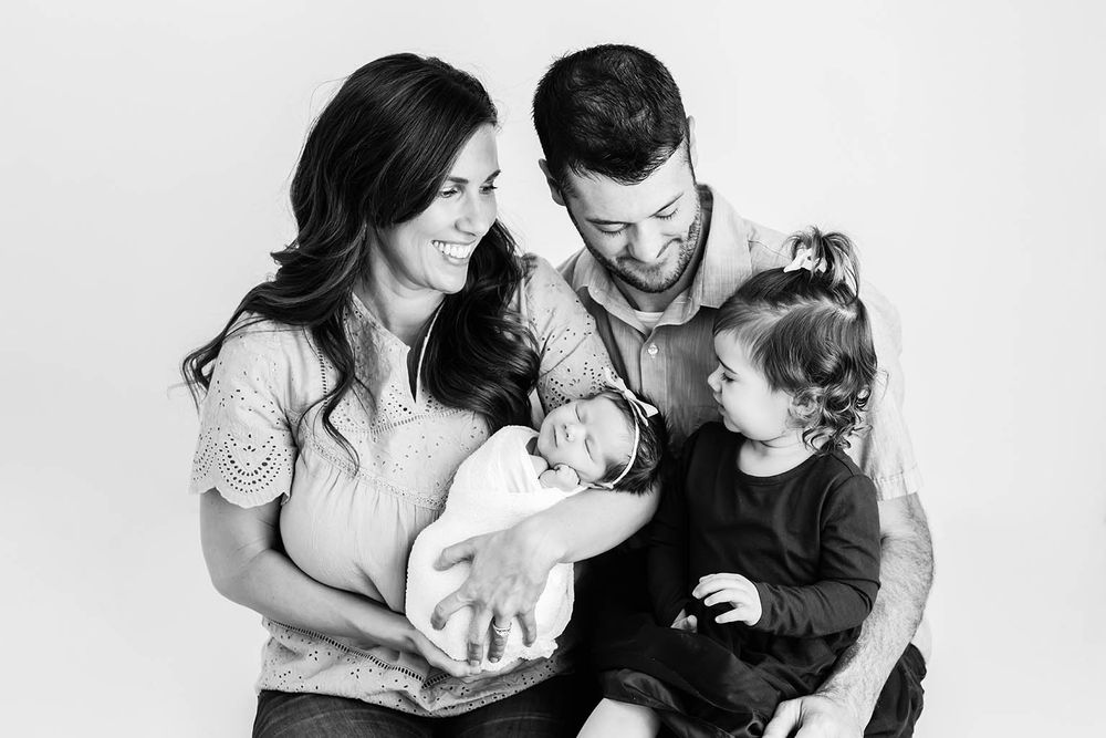 Beautiful black and white family portrait with parents holding newborn baby and smiling toddler daughter.