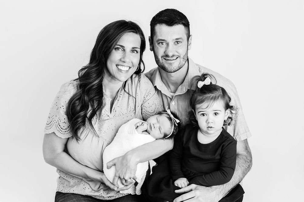 Black and white family portrait with parents holding newborn baby and toddler girl smiling.