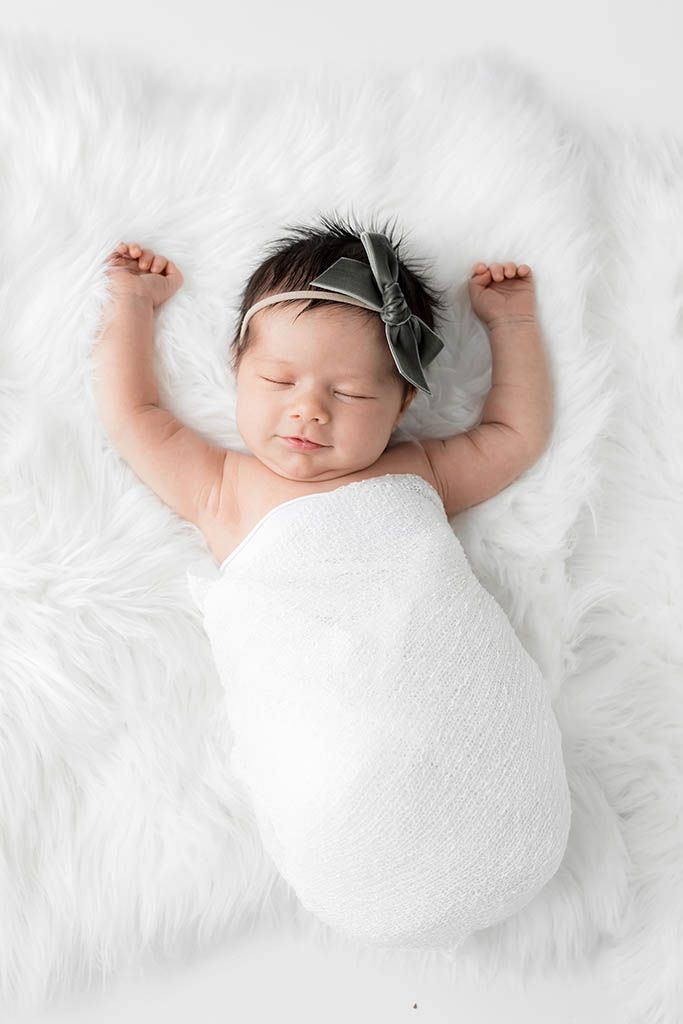 Sleeping newborn baby girl swaddled in white wrap with feather headband, arms raised on white fur.