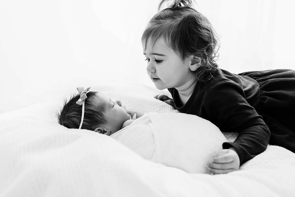 Toddler girl gazes lovingly at swaddled newborn sibling lying on white blanket.