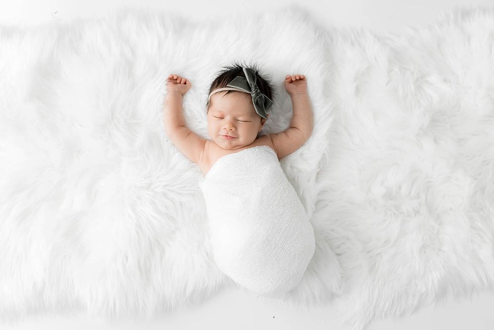 Newborn baby swaddled in white wrap with headband, sleeping peacefully on fluffy white fur rug.