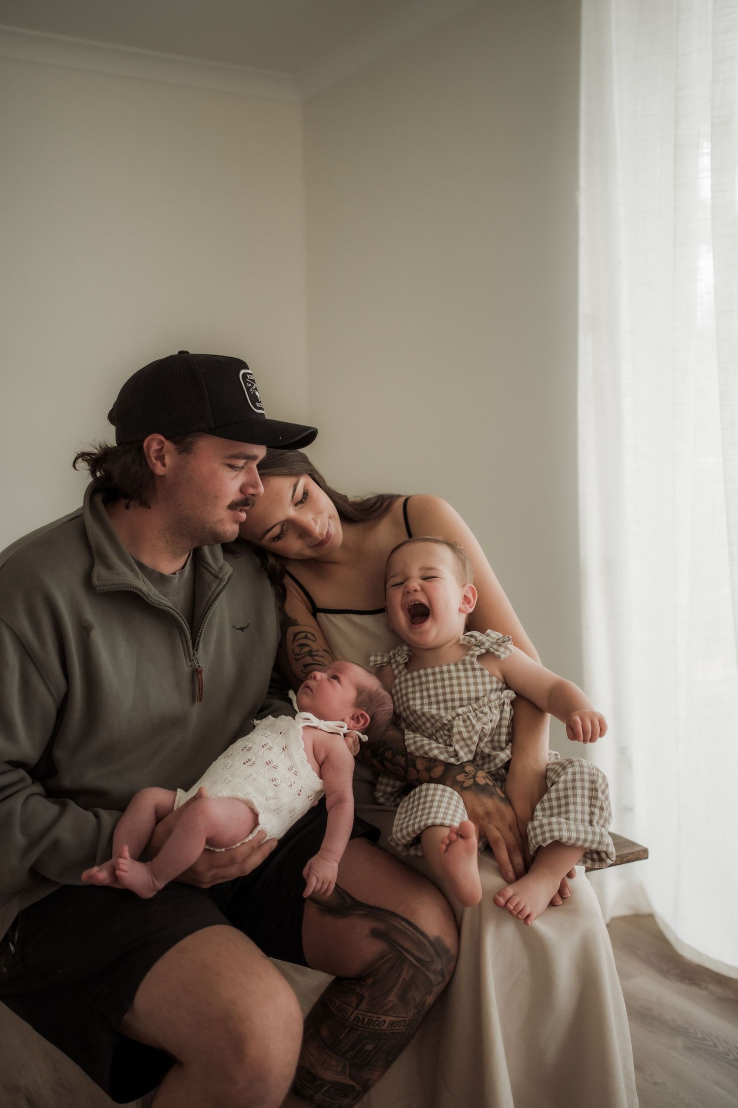 A young family sits together near a window, parents holding a newborn and a laughing toddler in a warm indoor setting in Buxton, Victoria