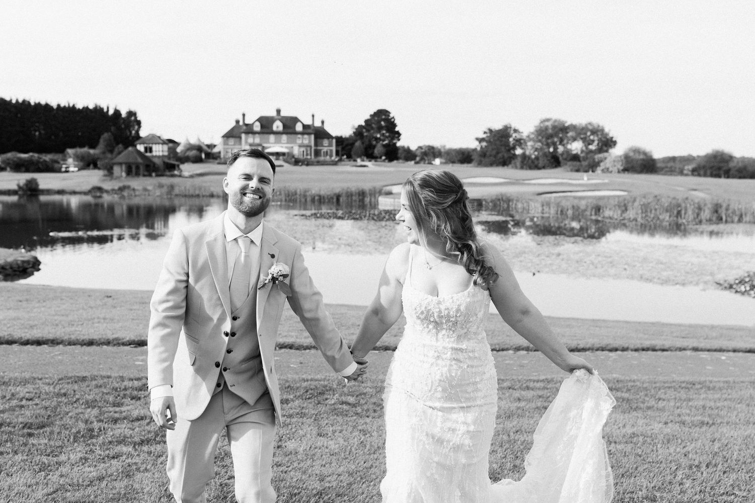 Black and white photo of a smiling groom in a suit holding hands with his bride in a lace wedding dress outdoors.