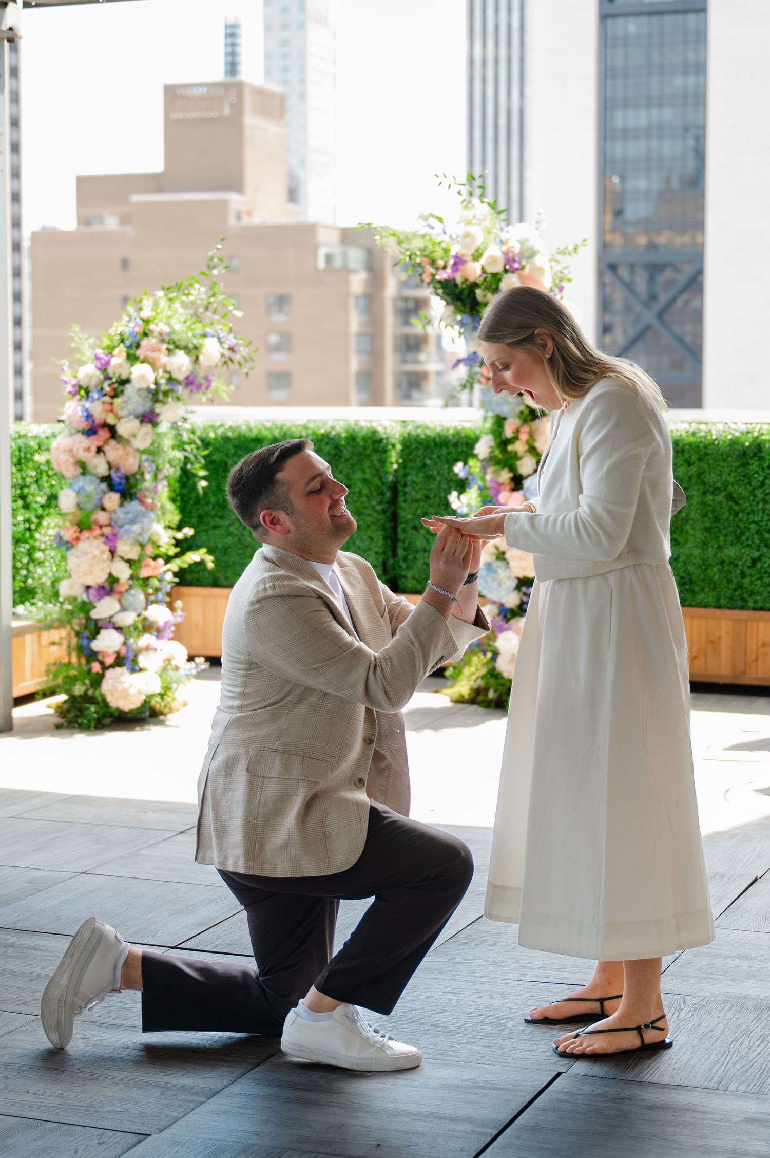 Man proposes on one knee to woman on a rooftop terrace in NYC at Le Jardin with floral arch and city skyline backdrop.