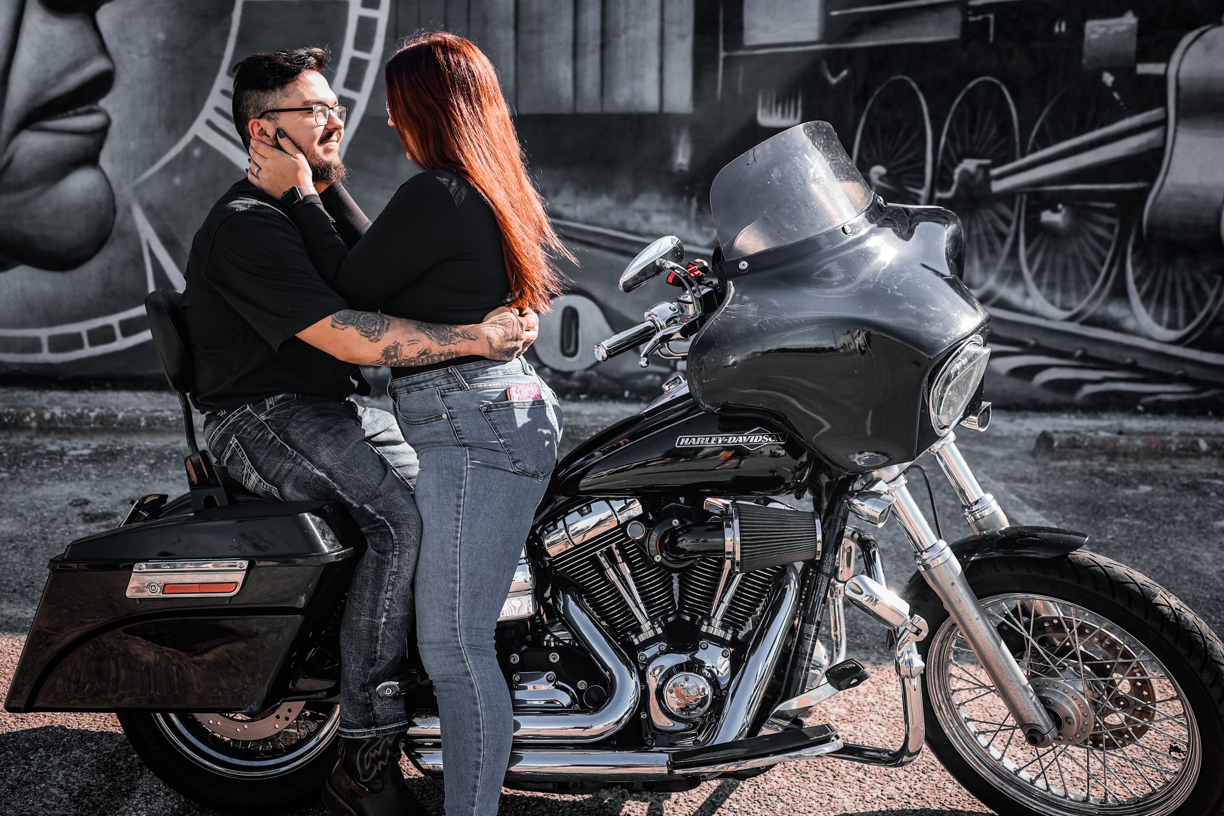 A couple shares a romantic moment sitting together on a Harley-Davidson motorcycle near a mural.