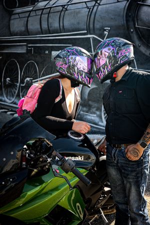 A tattooed couple in matching purple helmets lean in for a kiss beside a green Kawasaki motorcycle.