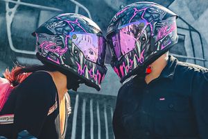 Couple sharing a kiss while wearing matching purple and pink graphic motorcycle helmets.