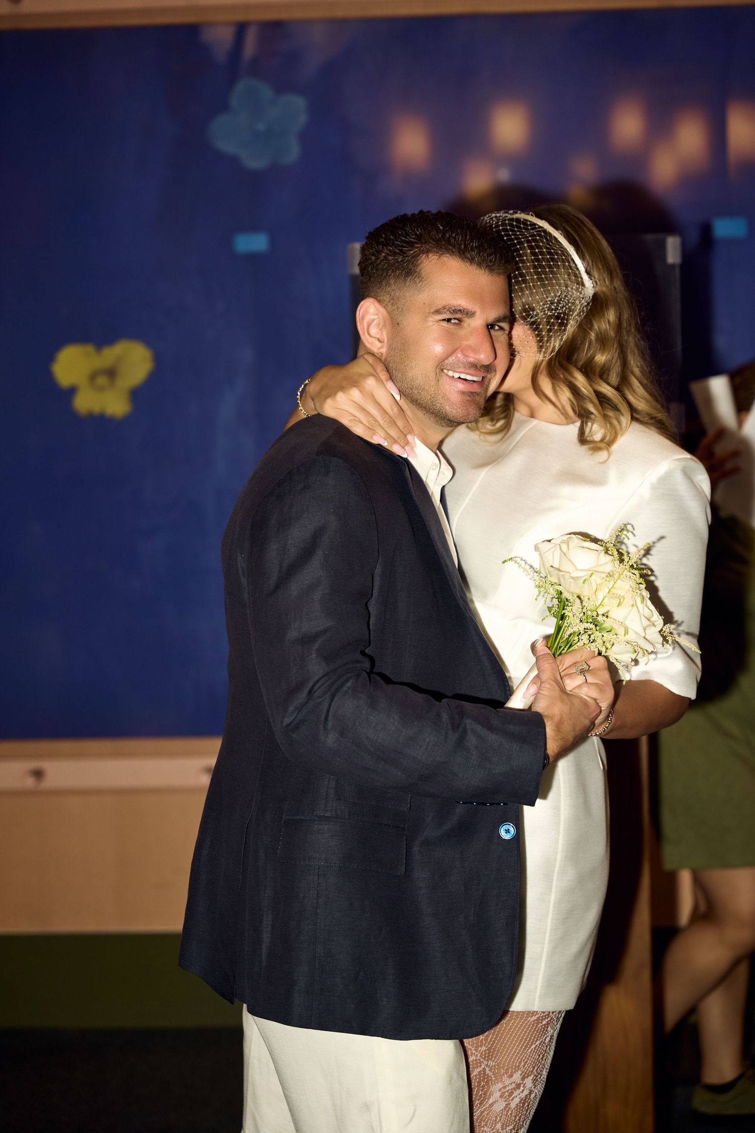 A smiling groom in a navy suit embraces his bride in a white dress holding a bouquet at their wedding reception.