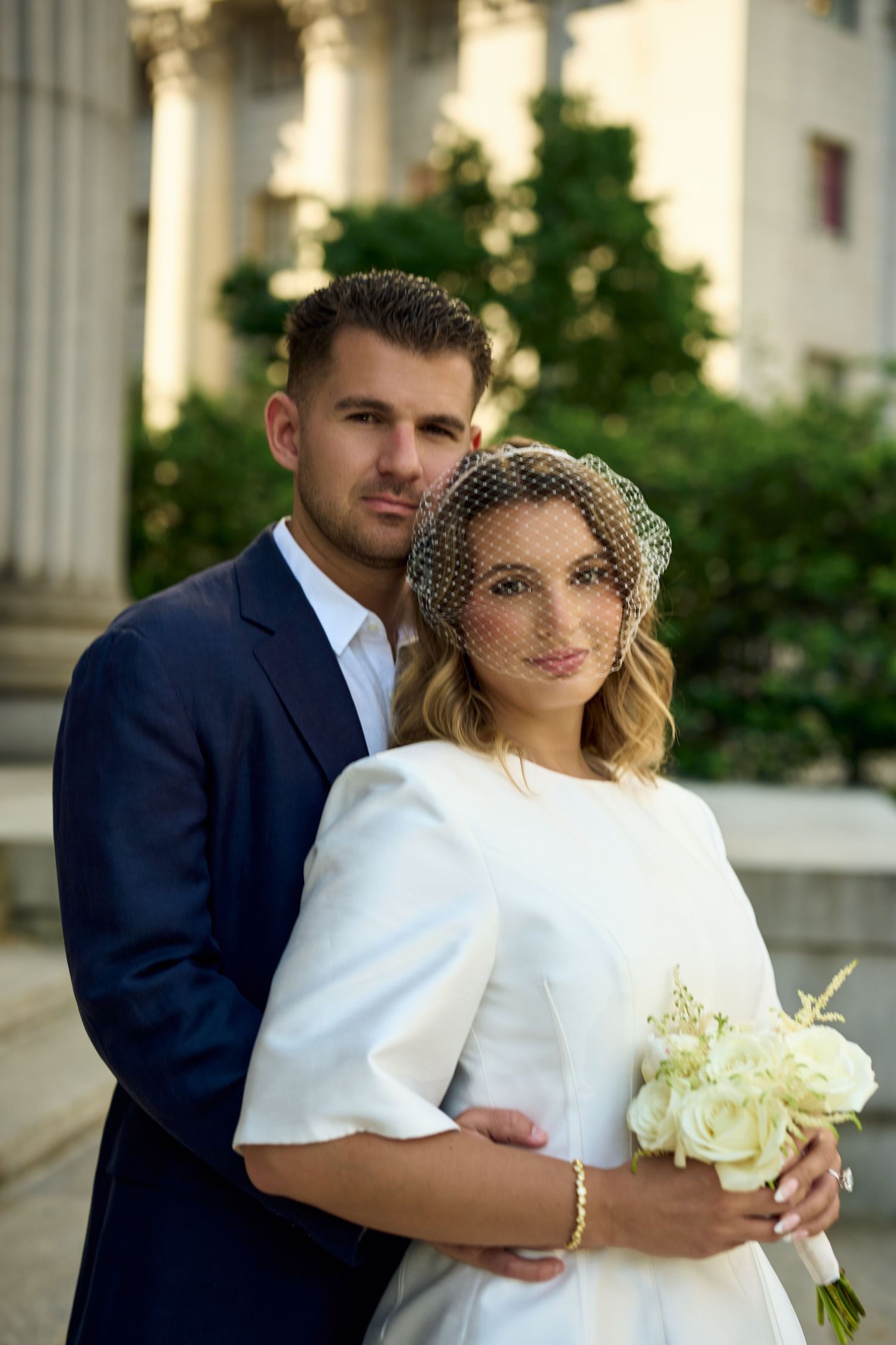 Bride in white dress with veil holding bouquet, groom in navy suit embracing her outdoors near columns.