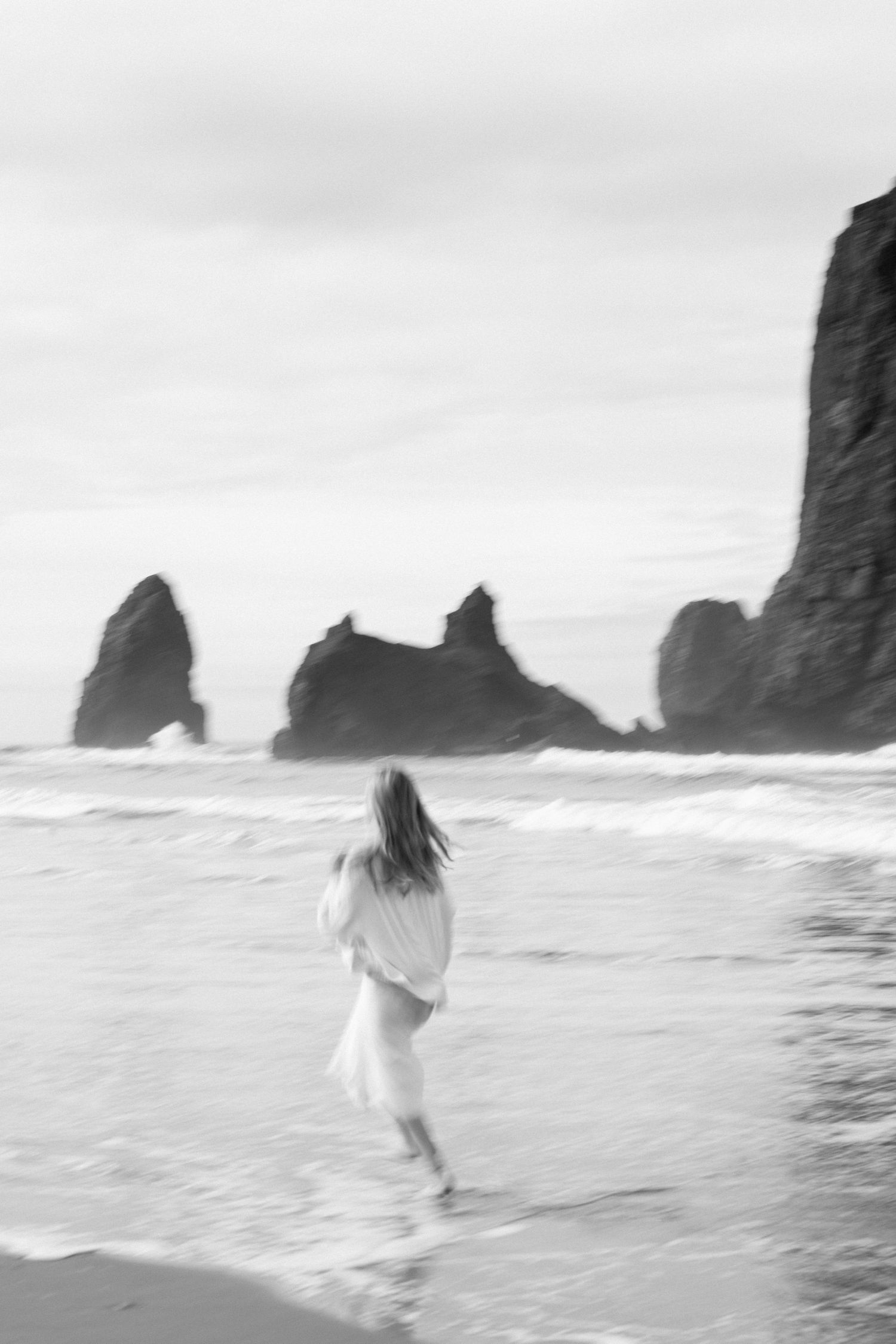 Black and white photo of a woman in a white dress walking along a rocky beach with dramatic sea stacks in the background.