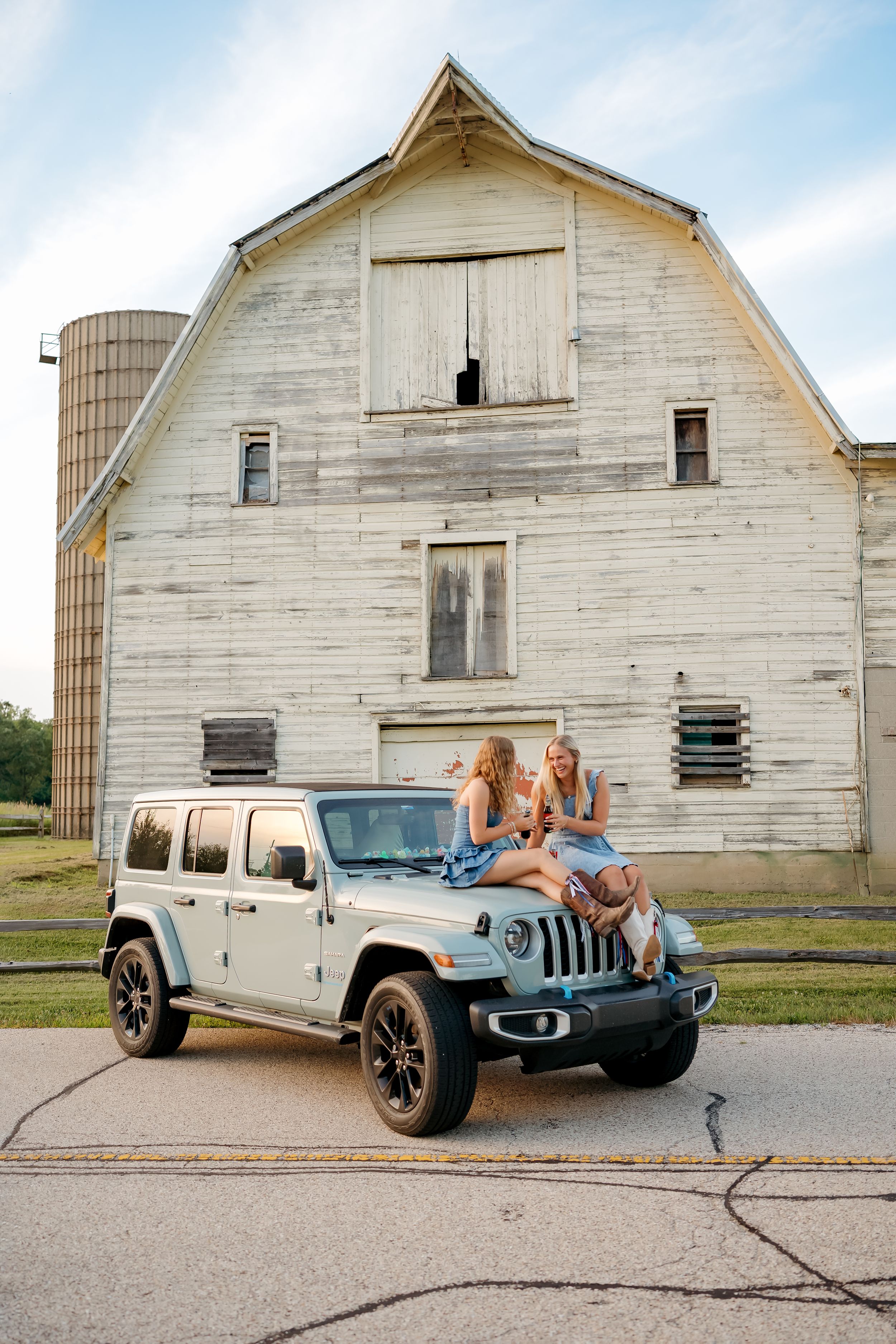 Two women sitting on a silver Jeep Wrangler hood in front of a rustic white barn with silo on a farm.