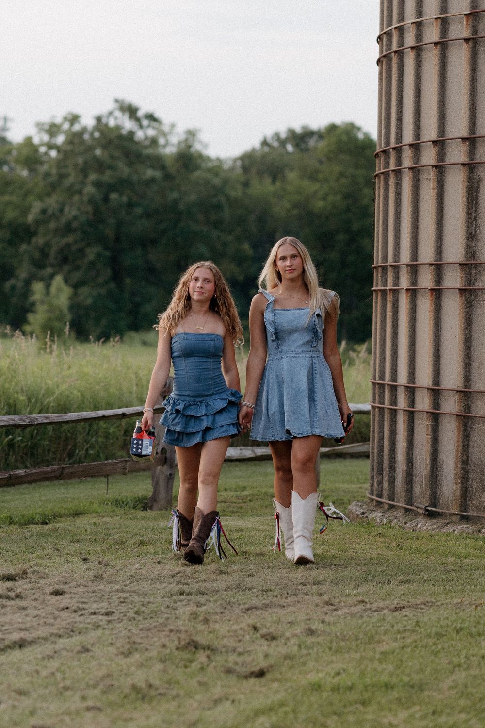 Two young women in denim dresses and cowboy boots walk outdoors near a silo surrounded by green fields.