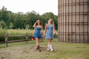 Two blonde women in matching blue denim dresses and cowboy boots walk hand-in-hand near a rustic farm silo.