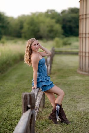 Young blonde woman in denim dress and brown cowboy boots posing against a rustic wooden fence in a green field.