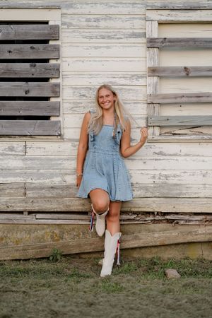 Smiling blonde woman in denim dress and white cowboy boots leaning against rustic wooden barn wall.