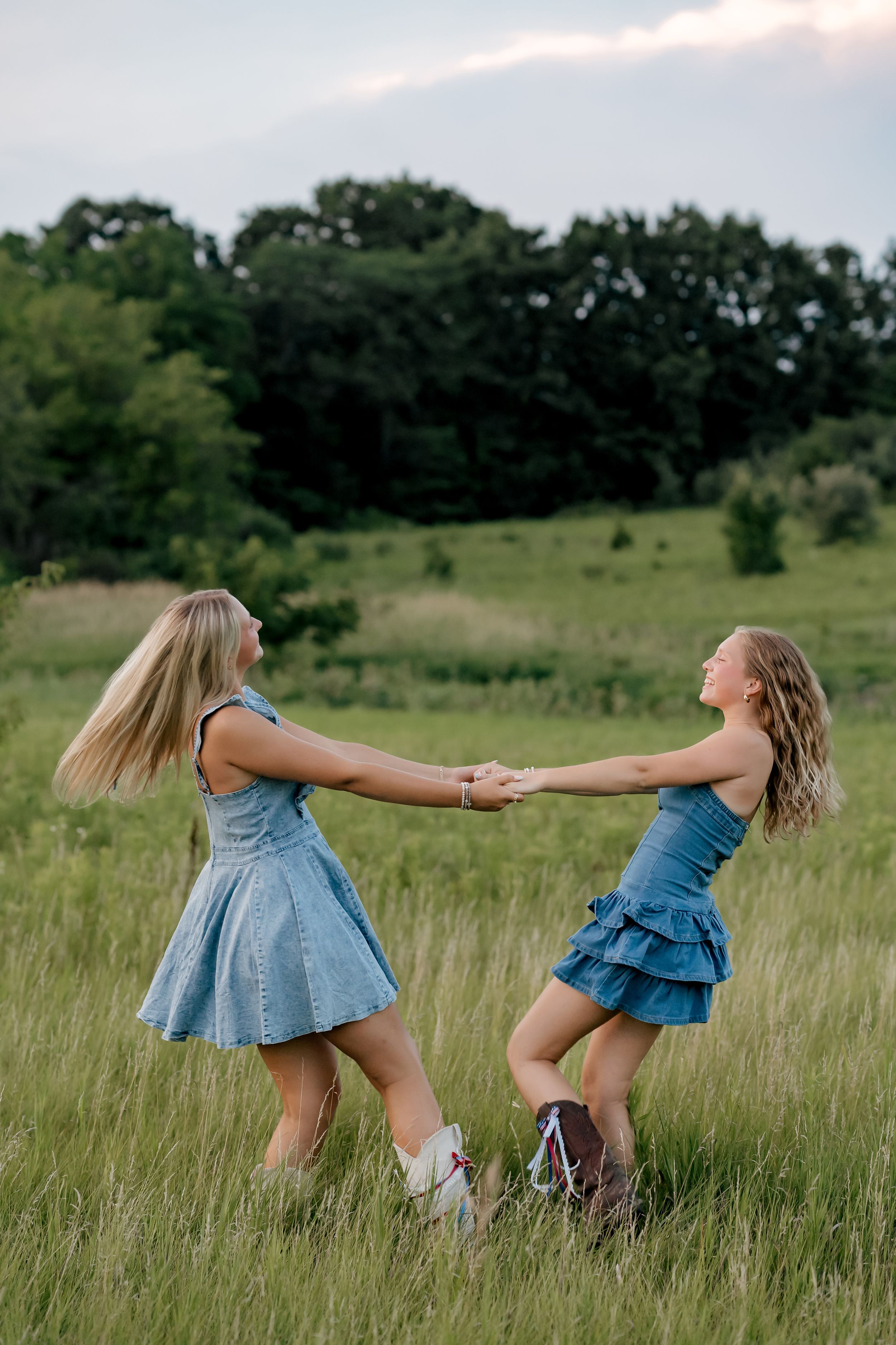 Two girls in denim dresses and cowboy boots spin holding hands in a green meadow.