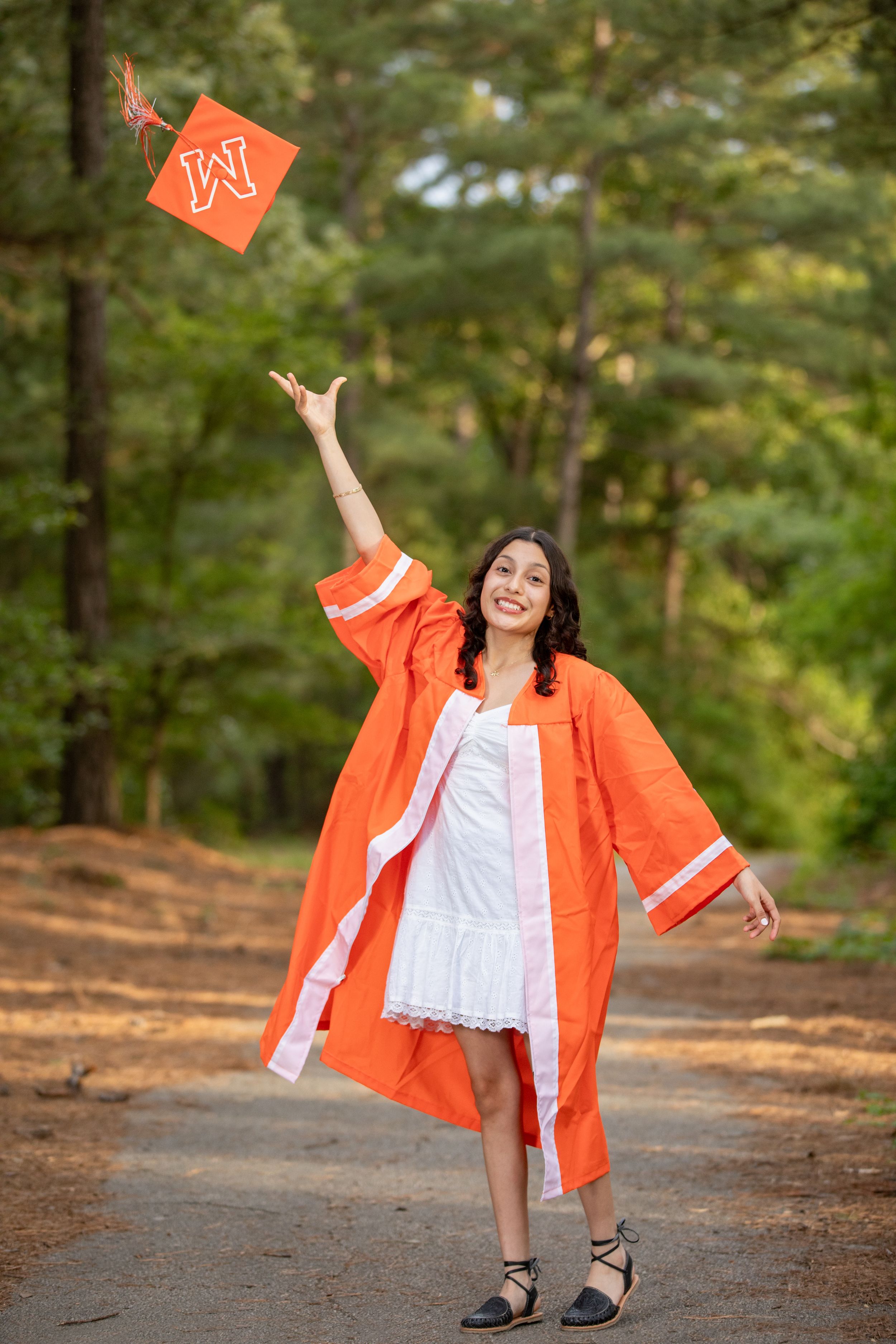 Graduate in orange regalia tossing graduation cap in celebratory pose on wooded path.