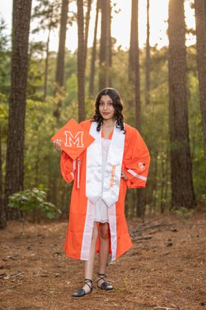 Graduate wearing bright orange graduation gown holds decorated cap on forest trail.