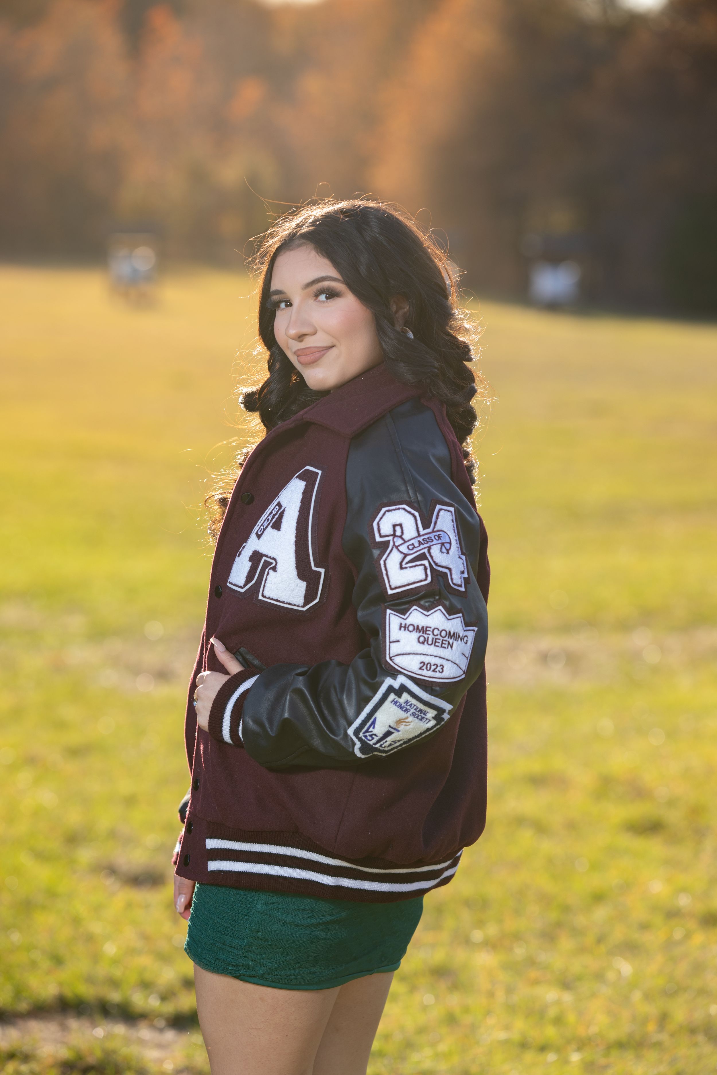 Student in varsity letterman jacket poses outdoors during golden sunset.