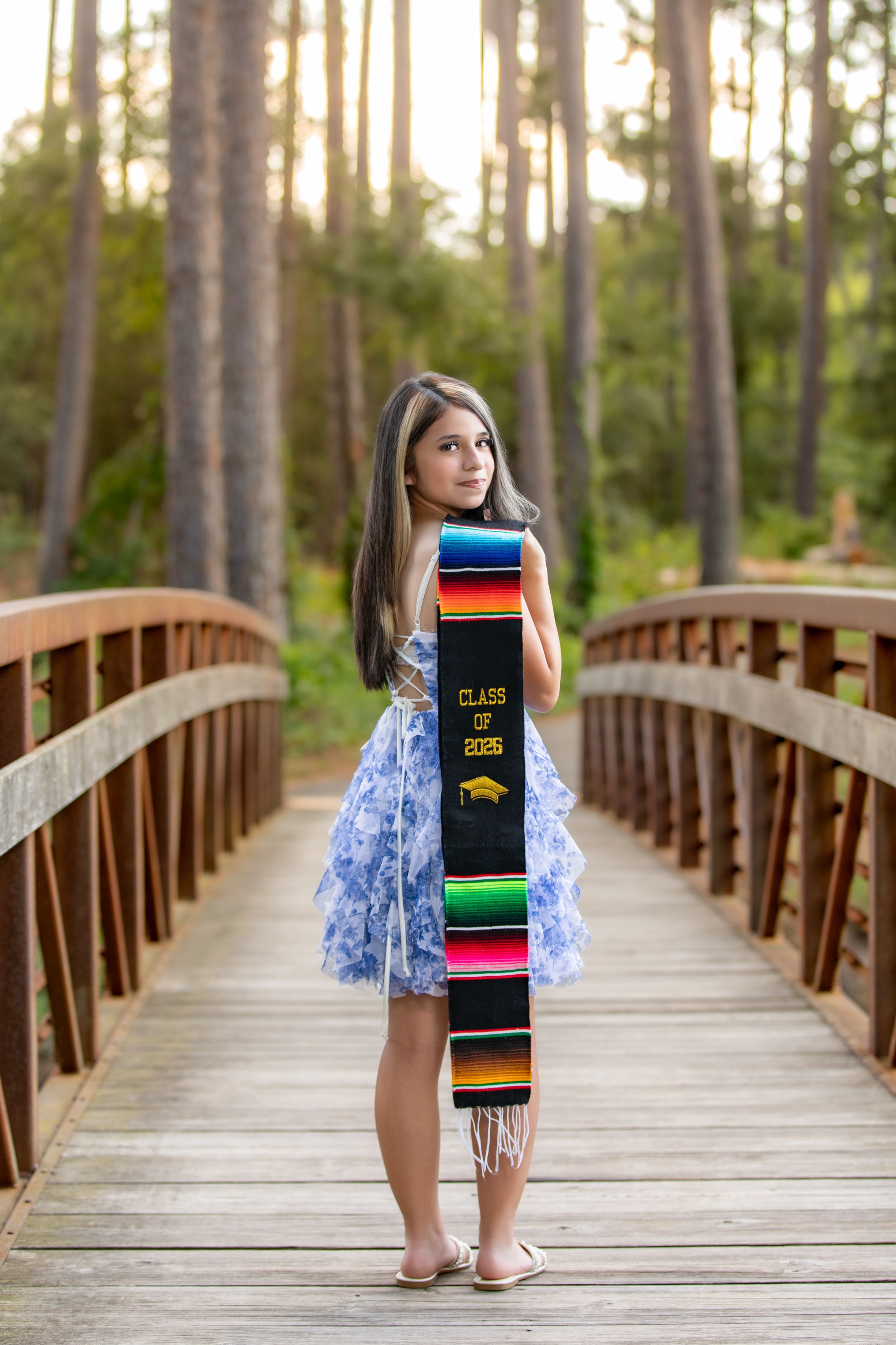 Young woman in blue dress holding a colorful Class of 2025 graduation stole on a wooden bridge in a forest.