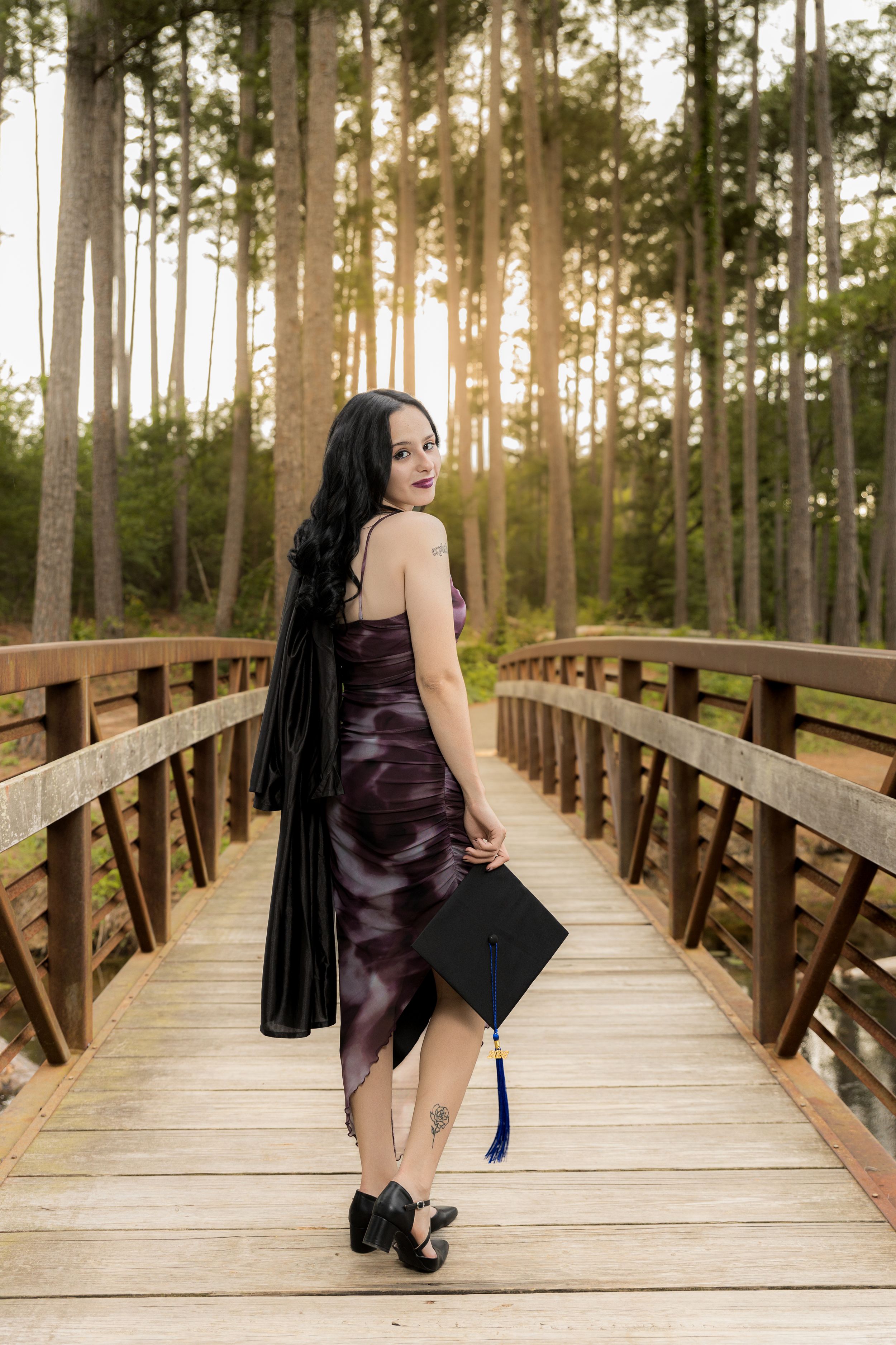 Young woman in purple dress holding graduation cap on wooden bridge surrounded by tall pine trees at sunset.