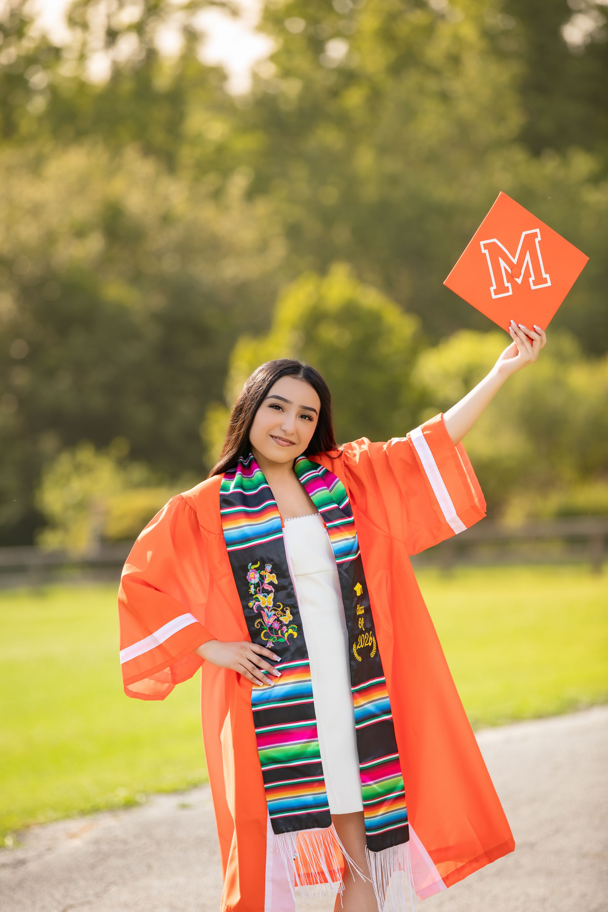 Young woman in orange graduation gown holds cap aloft, wearing colorful serape stole, posing outdoors on a sunny day.