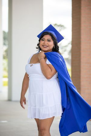Graduate in flowing white dress and royal blue academic regalia poses confidently.