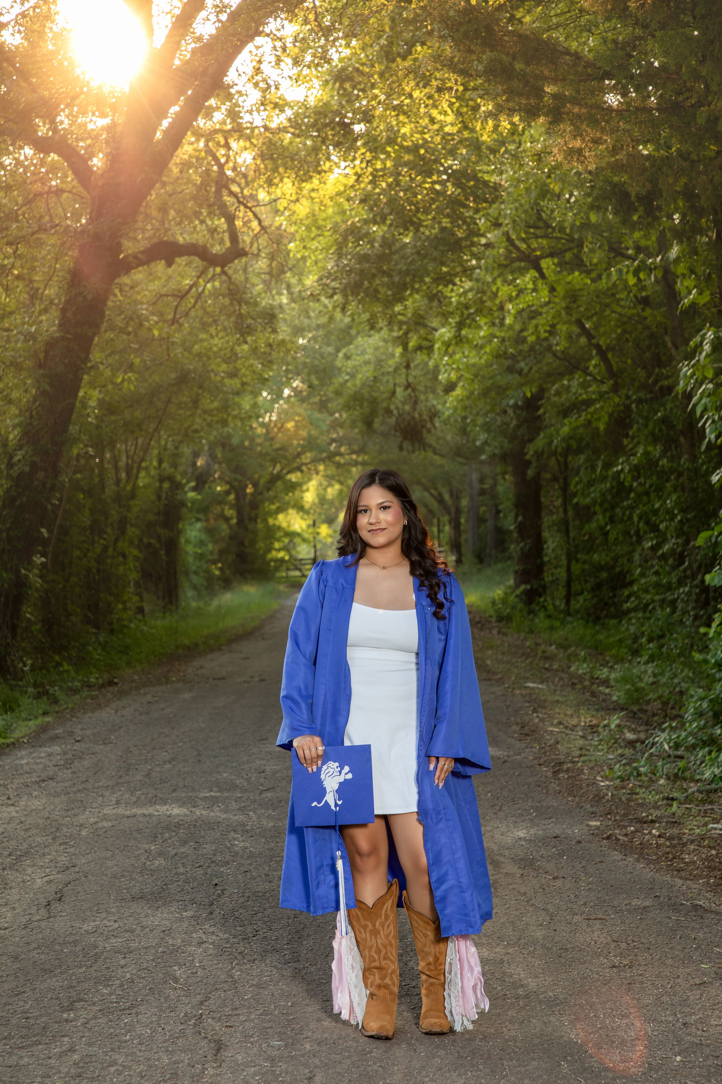 Young woman in blue graduation gown and cowboy boots holding cap on a sunlit forest path.
