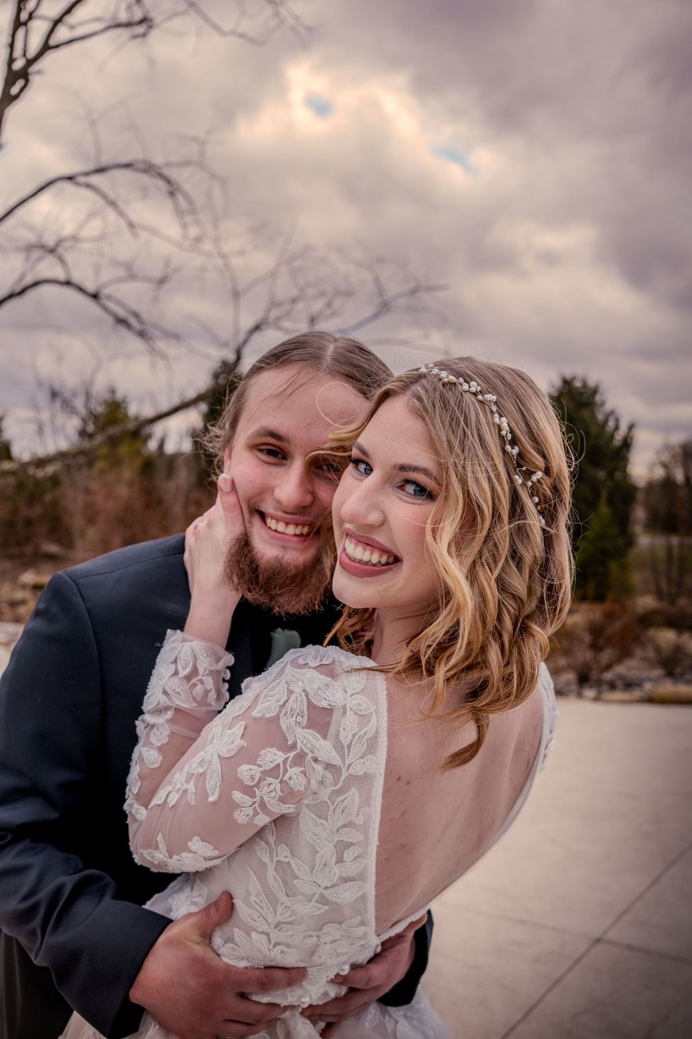 Smiling bride in lace gown with floral hair accessory embraces groom in navy suit outdoors under cloudy sky.