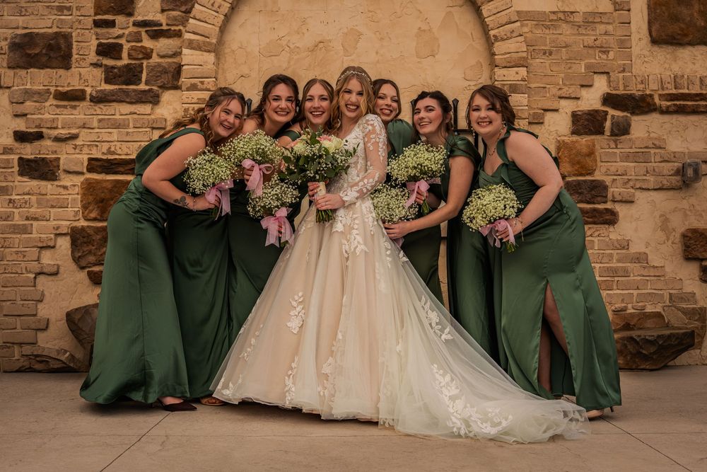 Bride in lace gown poses with six bridesmaids in emerald green dresses holding pink bouquets before stone wall.