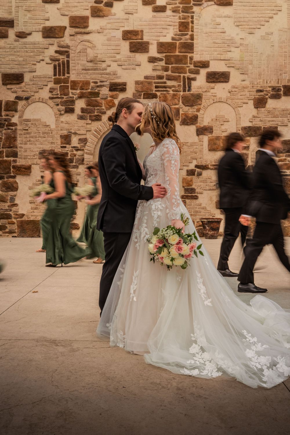 Bride and groom share a kiss in front of a stone wall, with blurred wedding party members in green and black attire.