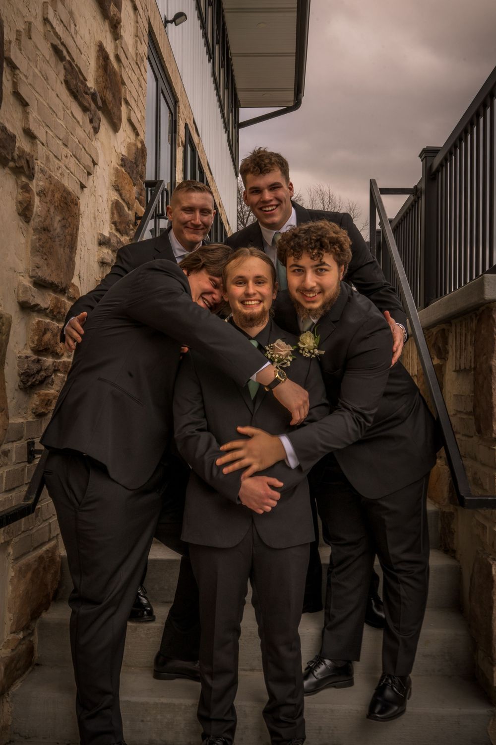 Group of five young men in dark suits hugging and smiling on outdoor steps at a formal event.