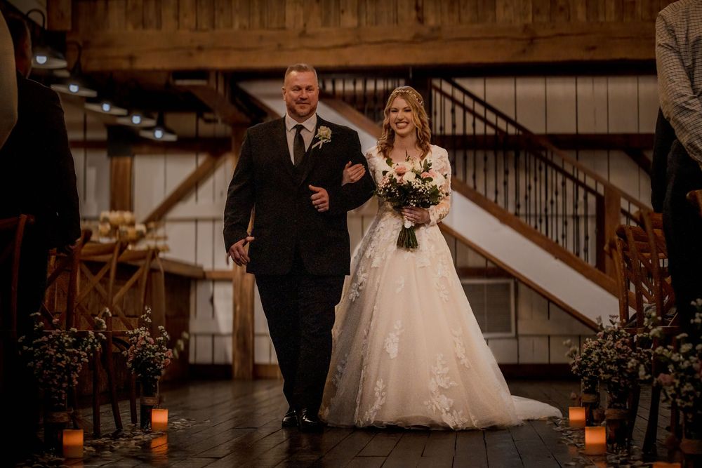 Bride in floral lace gown walks down aisle with escort in rustic barn wedding venue with candlelit path.