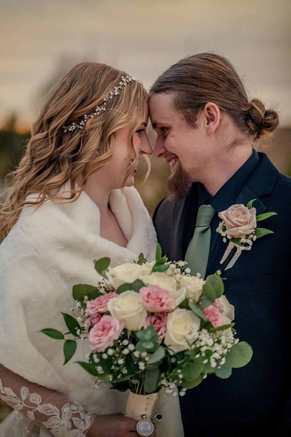 Bride and groom touching foreheads, smiling, holding pink and white rose bouquet, outdoor winter wedding portrait.