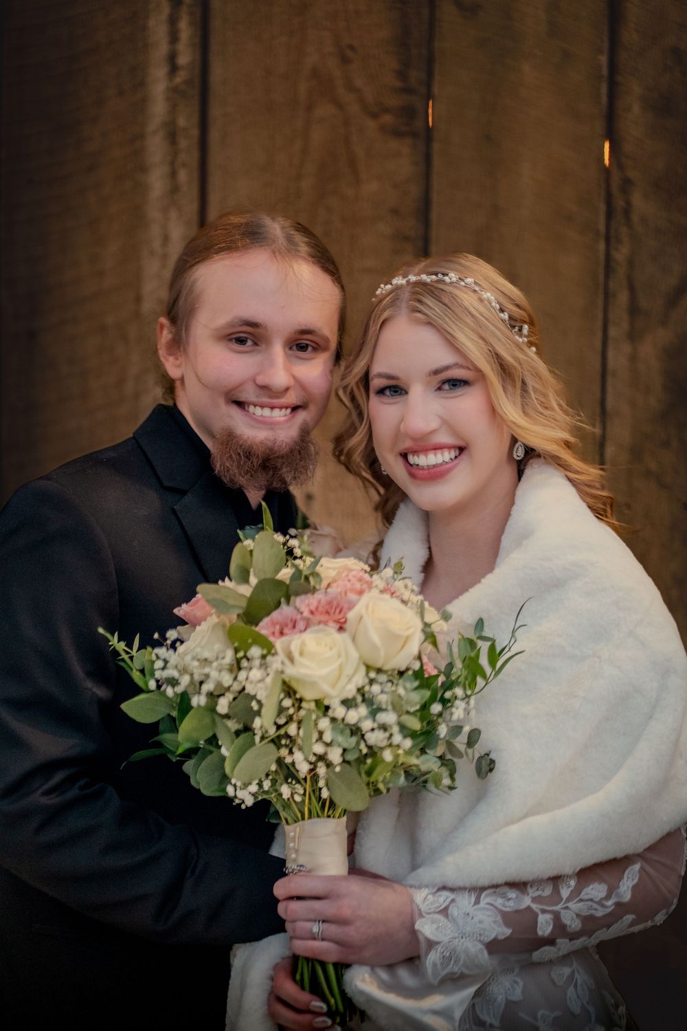 Smiling bride and groom pose together, bride holding pink and white floral bouquet, wearing white wrap and floral headpiece.
